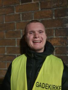 Frivillig fra Gadekirke i Randers smilende i refleksvest under gadeevangelisation foran murstensvæg.